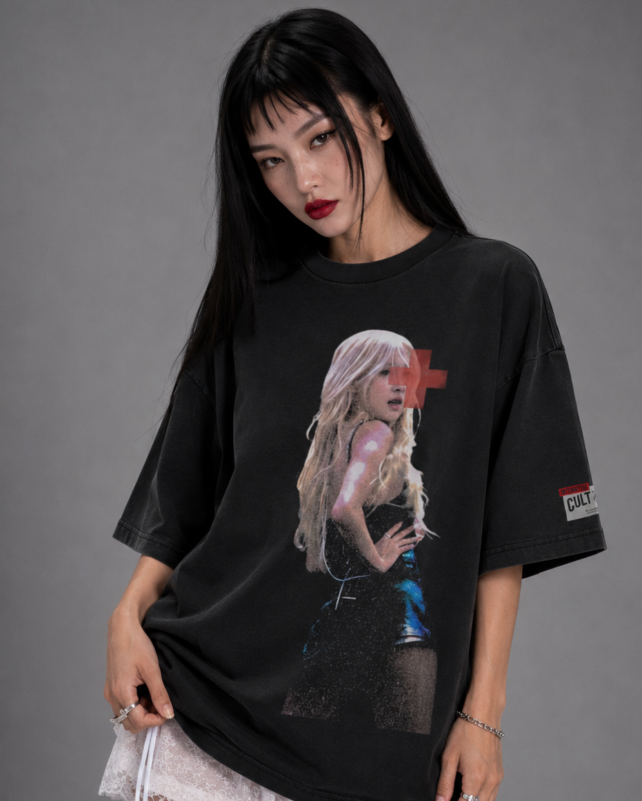 A woman modeling an oversized black graphic t-shirt with a large print of a blonde woman on the front, paired with a white lace hem mini skirt, against a solid gray studio background.